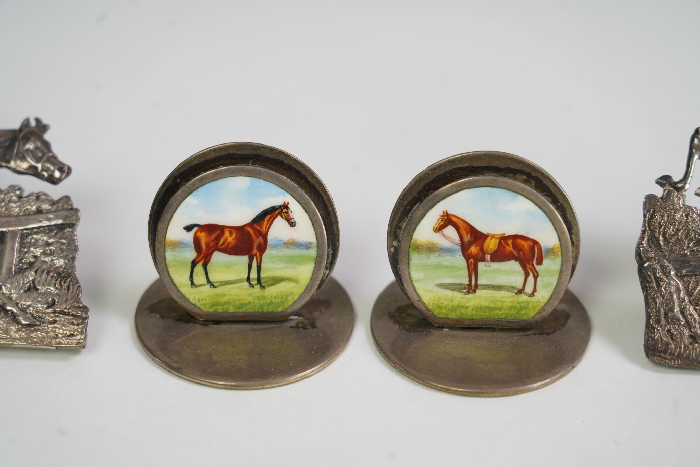A pair of late Victorian silver menu holders, each modelled as a horse and rider leaping a fence, by Samuel Jacob, London 1894/5, height 66mm, together with a pair of Edwardian silver and enamel menu holders, each decora
